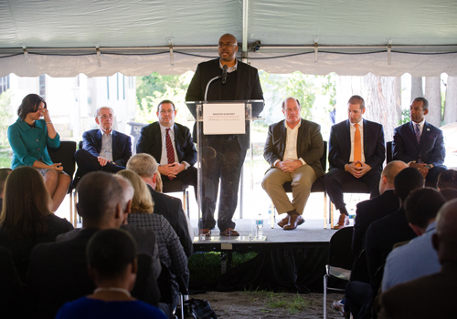 Man speaking at podium in white tent with audience in front of him and others seated in a line behind him