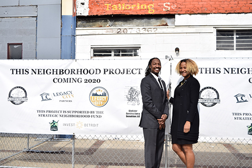 Two people smiling in front of a sign saying "This Neighborhood Project, Coming 2020"